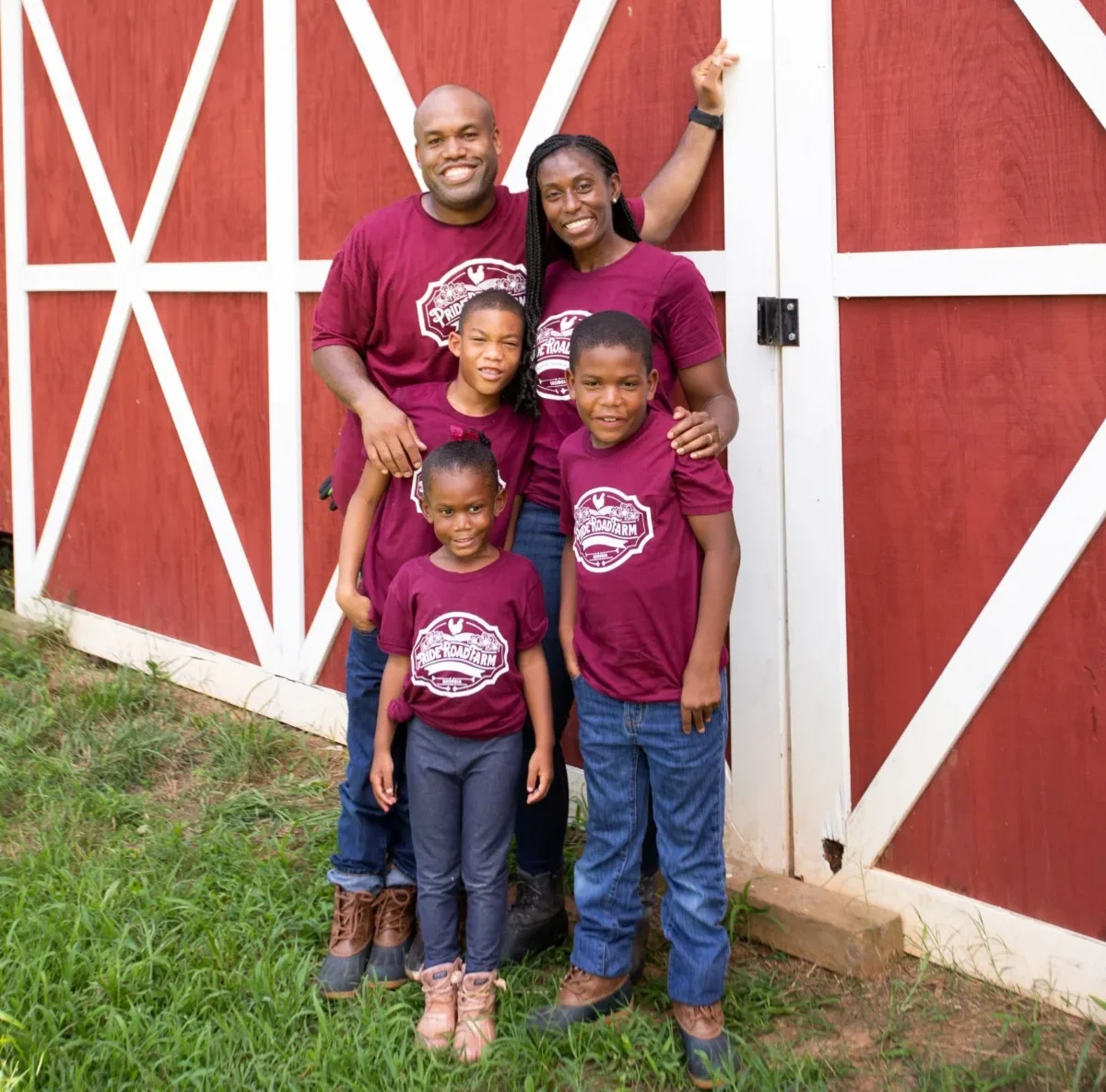 A father, mother, and three childen wearing red shirts stand in front of a red barn.