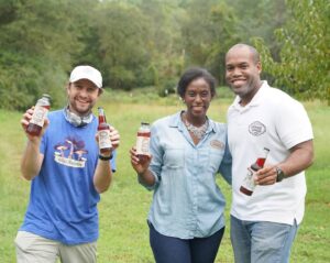 Three people stand in a field holding bottles of a red drink (hibiscus tea)