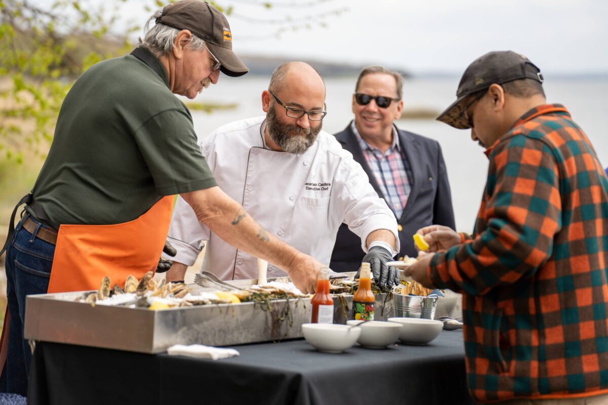 Four men - one in an orange apron, one in a chef's coat, gather around an outdoor raw bar. 