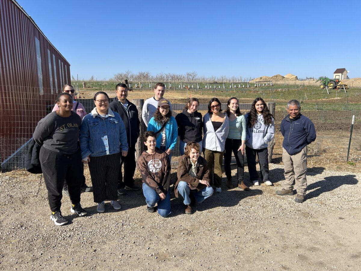 A group of 13 people stands in front of a barb with a field behind them. 