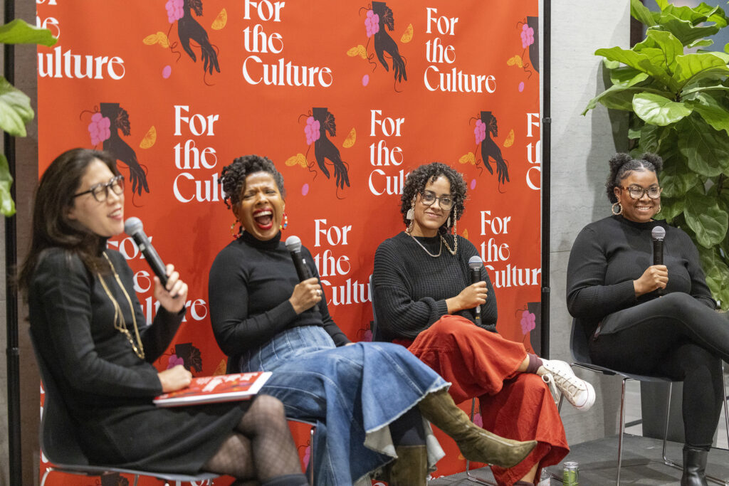 A group of women seated in a row for a panel discussion in front of a bright orange graphic background. 