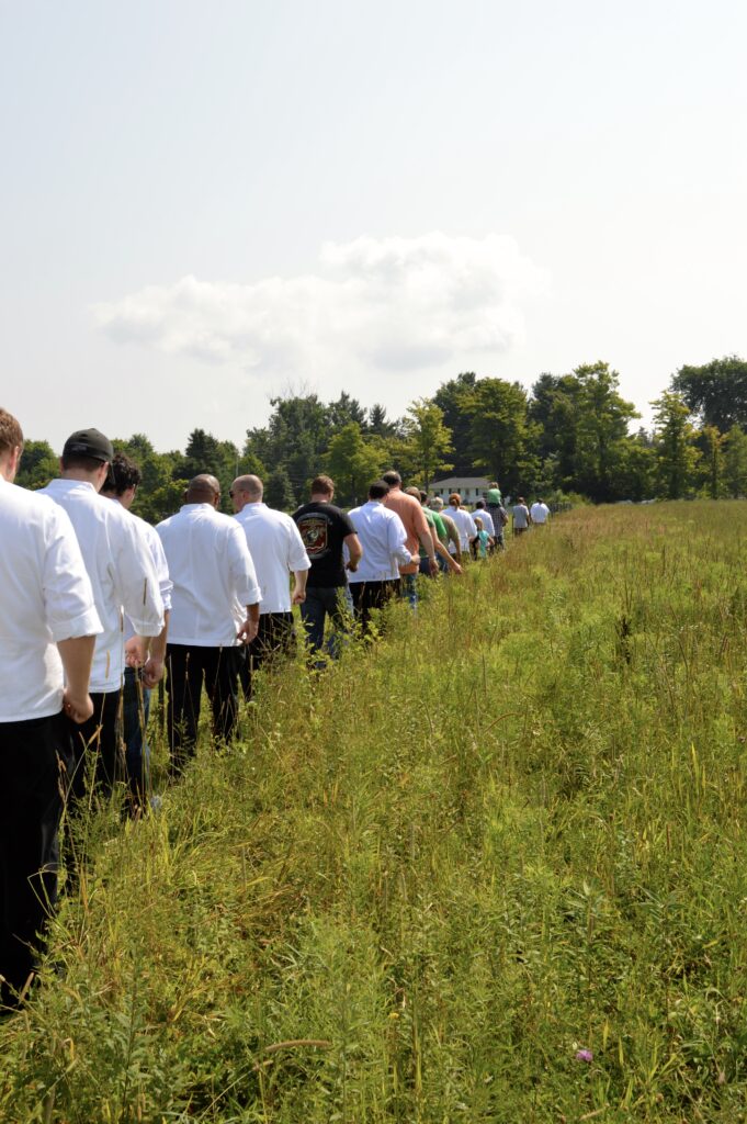 A group of people wearing white chefs' coats walk single file through a field. 