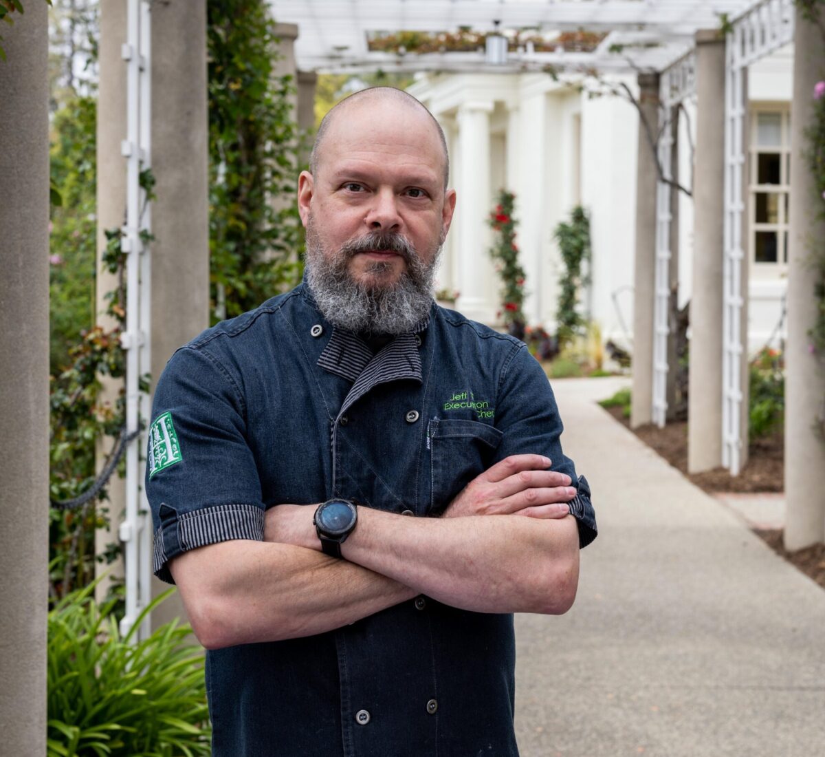 A man in a short-sleeved chef coat stands in front of a long portico 