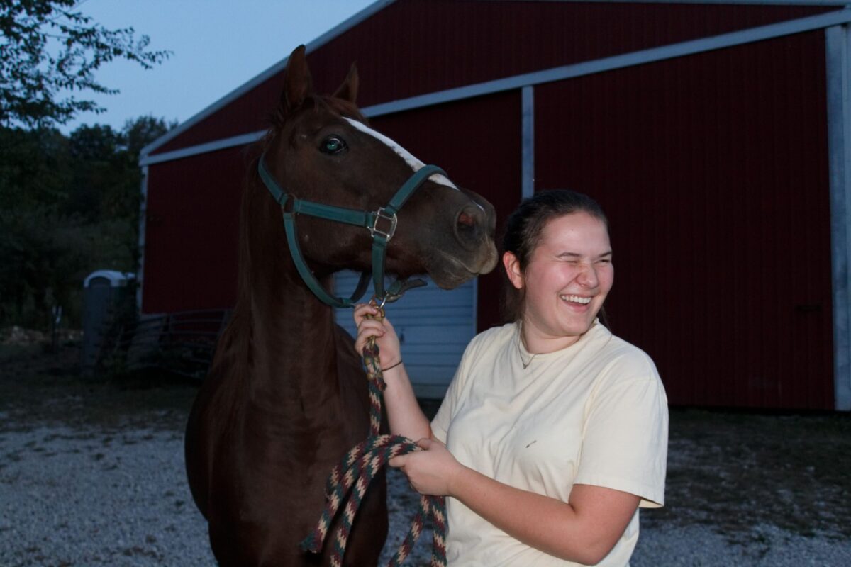 A young woman holds a horse's lead while it tries to lick her face.