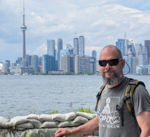 A man in a t-shirt and sunglasses stands in front of Toronto's skyline