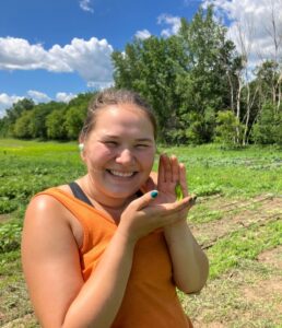 A young woman in an orange tank top holds up a pepper while standing in a field. 