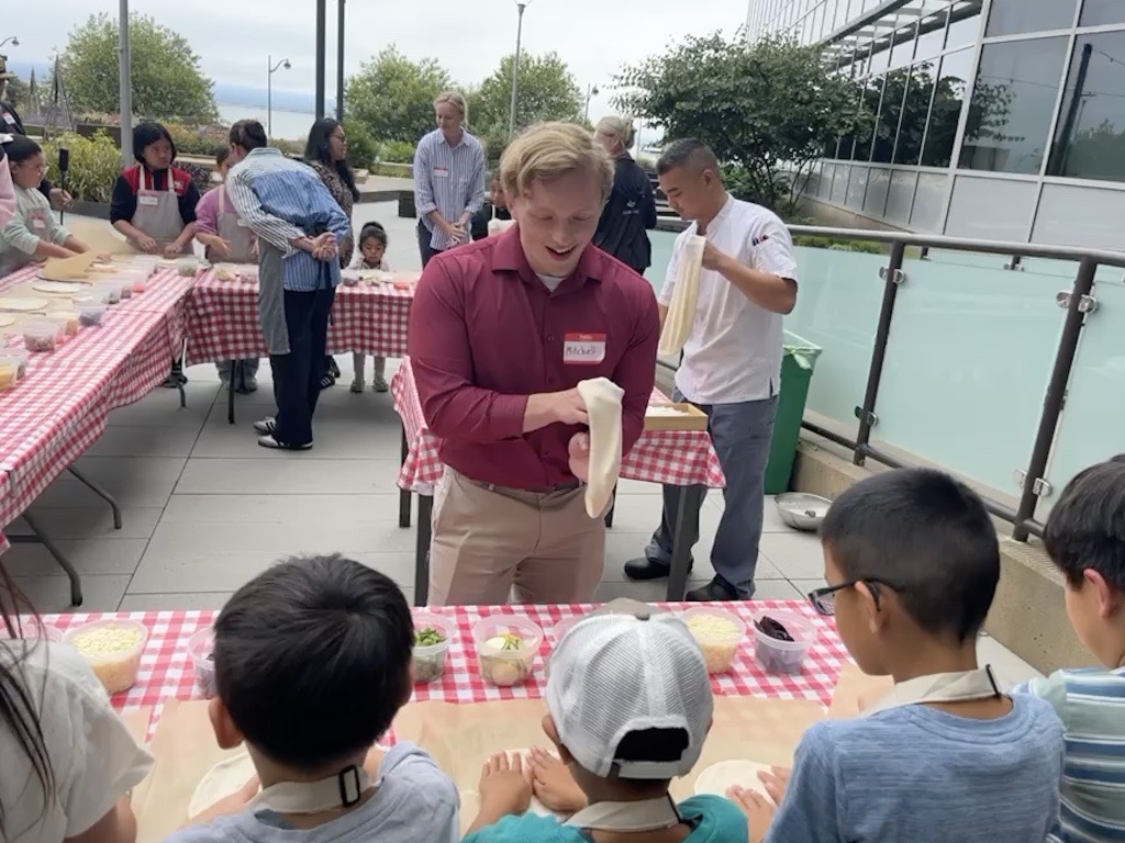 A young man wearing a dark red shift demonstrates tossing pizza dough for a group of young children as part of Bon Appetit's Healthy Kids program. 