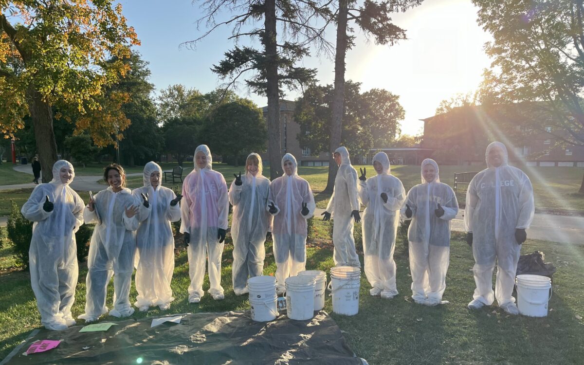 A group of young people in head-to-toe protective gear stand in a semi-circle with three buckets of waste in front of them.