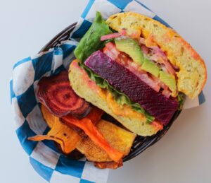 A crossways view of a burger made with a thick slice of beets, with avocadoes, pickled onions, spicy may, and a pumpkin bun.