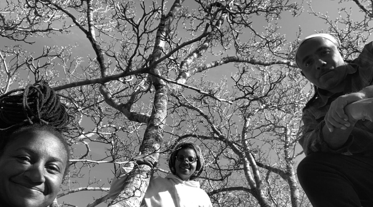 A black and white photo of three people - two women and one man - standing around a tree and looking down at the camera. 