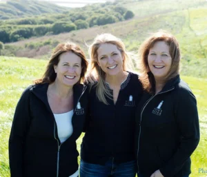 Three women stand together with rolling green hills behind them.